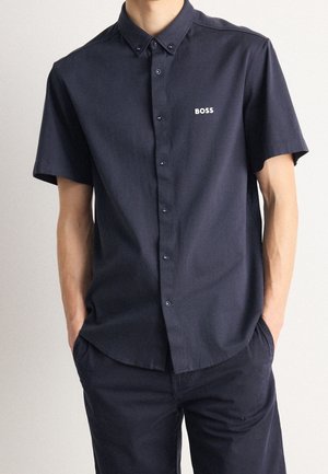 Mężczyzna w ciemnoniebieskiej krótkiej koszuli zapinanej na guziki z logo "BOSS" na piersi, ręce w kieszeniach dopasowanych ciemnoniebieskich spodni.