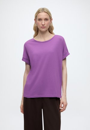 Γυναίκα με ανοιχτόξανθα μαλλιά, φορώντας χρυσούς κρίκους στα αυτιά, μια φαρδιά μωβ μπλούζα με κοντά μανίκια και μαύρο παντελόνι, μπροστά από ένα απλό φόντο.