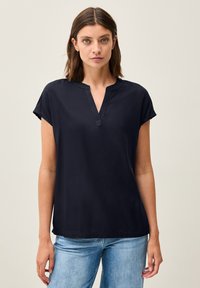 Blusa blu navy a maniche corte con scollatura a V e texture liscia, abbinata a jeans di denim blu chiaro. Fit semplice e rilassato.