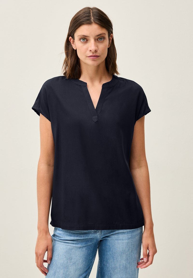 Blusa blu navy a maniche corte con scollatura a V e texture liscia, abbinata a jeans di denim blu chiaro. Fit semplice e rilassato.