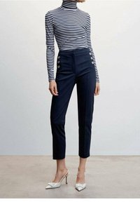 Pantalons taille haute marine avec des accents de boutons dorés ; associés à un col roulé rayé noir et blanc et des mules blanches à talons.