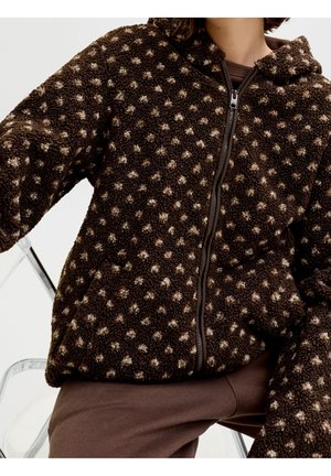 Person trägt eine braune Fleecejacke mit Reißverschluss und hellbraunen Polka-Punkten, sitzt auf einem transparenten Stuhl.