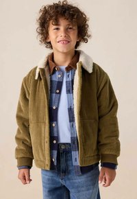 Veste en velours côtelé vert olive avec un col en fausse fourrure blanche, dotée d'une texture côtelée, de poches avant et accompagnée de chemises à carreaux bleus superposées et d'une chemise bleu clair.