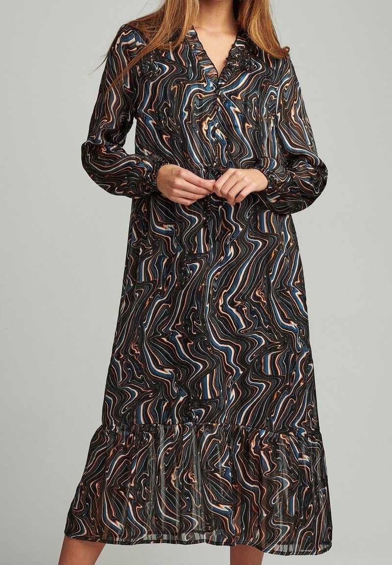 Femme portant une robe à manches longues, longueur genou, avec un motif tourbillonnant multicolore dans des tons de bleu, marron, noir et beige.