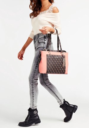 Kvinde iført off-shoulder top og grå jeans gående med sorte støvler og bærende en pink og mønstret Guess-taske på armen.