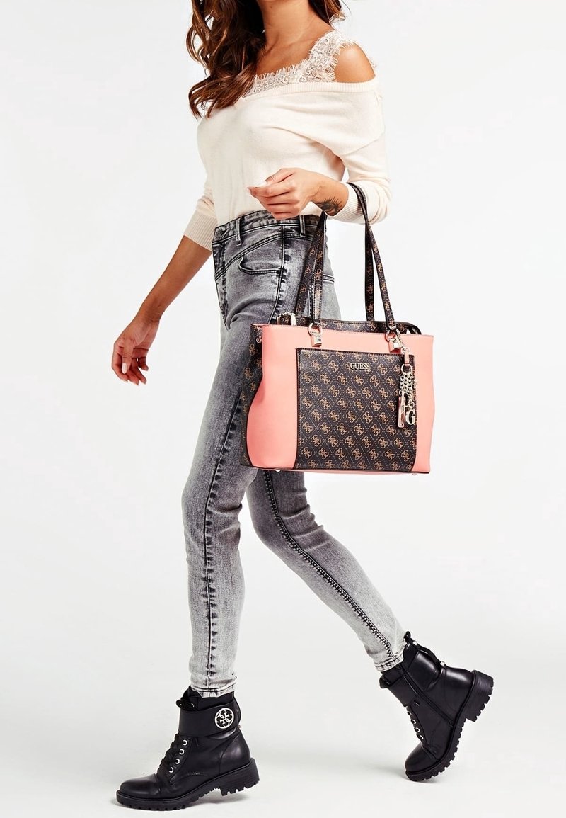 Femme portant un top épaules dénudées et un jean gris, marchant avec des bottes noires et portant un sac cabas Guess rose à motif sur le bras.