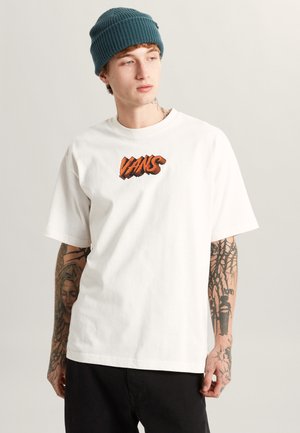 T-shirt bianco in cotone con maniche corte e collo rotondo, con un logo arancione "VANS" sul petto. Pantaloni neri e un berretto teal.