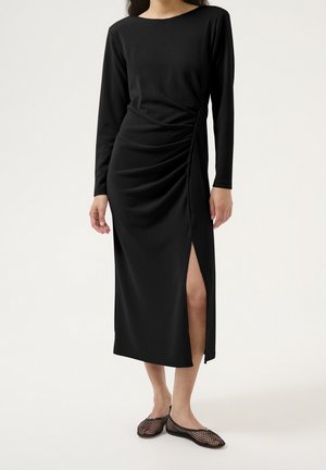 Femme portant une longue robe noire avec des fronces sur les côtés et une fente haute sur la cuisse, associée à des chaussures plates en résille noire.