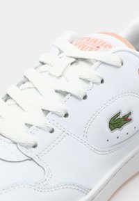 Fehér bőr sneaker, kerek orral, kontrasztos zöld krokodillogóval és texturált fehér fűzővel. A nyelvén rózsaszín akcentussal rendelkezik.