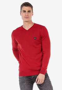 Roter Pullover mit langen Ärmeln und V-Ausschnitt. Verfügt über ein kleines schwarzes Logo-Patch auf der Brust sowie gerippte Bündchen und Saum. Strukturierter Stoff.