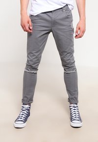 Šedé denimové slim-fit kalhoty se zdrsněnými detaily na kolenou, ohrnutými nohavicemi a hladkým povrchem. Nosič má na sobě navy modré vysoké tenisky.