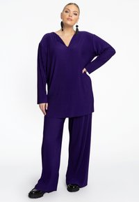 Paarse, geribbelde loungewear set met een langmouwtop met V-hals en broek met wijde pijpen, beide met een losse fit. Zwarte platform schoenen.
