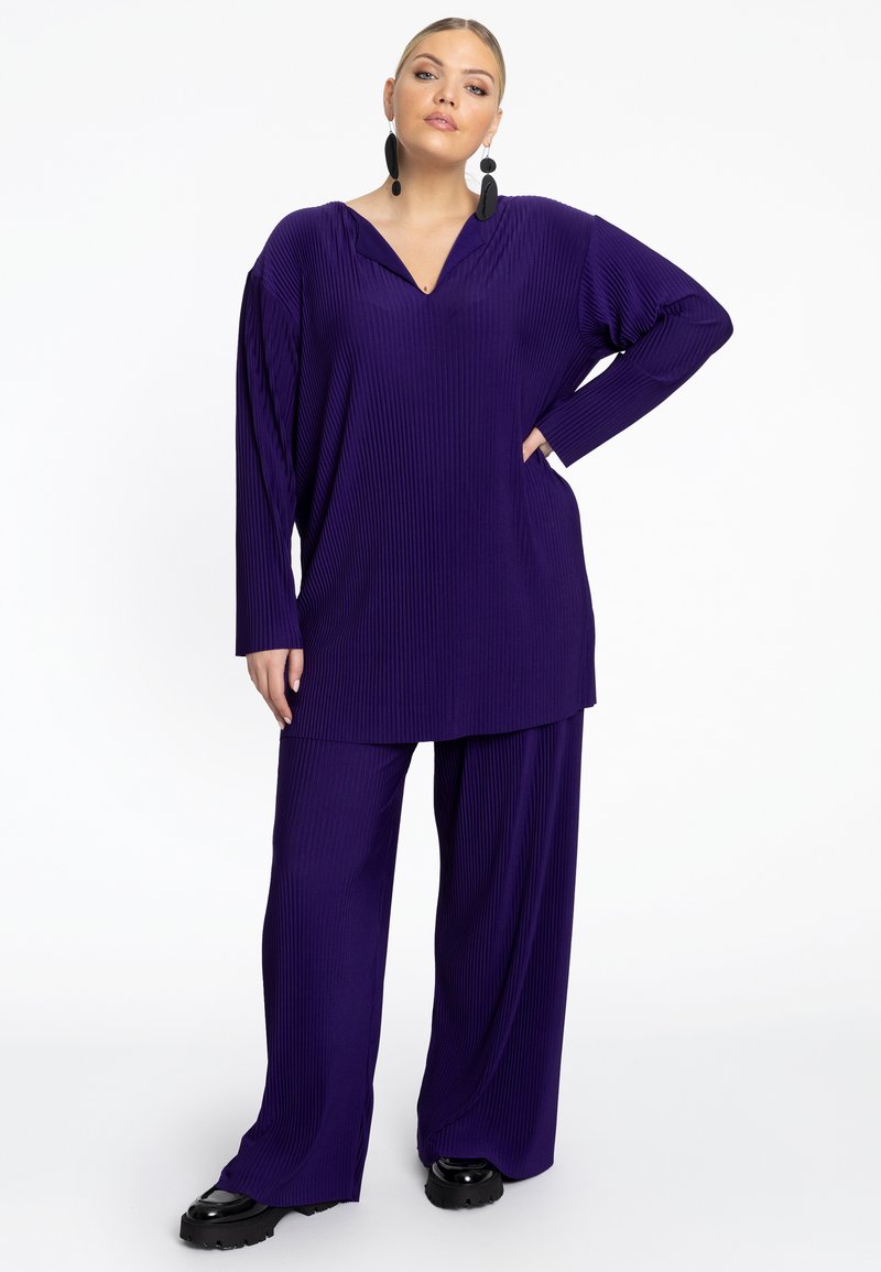 Paarse, geribbelde loungewear set met een langmouwtop met V-hals en broek met wijde pijpen, beide met een losse fit. Zwarte platform schoenen.