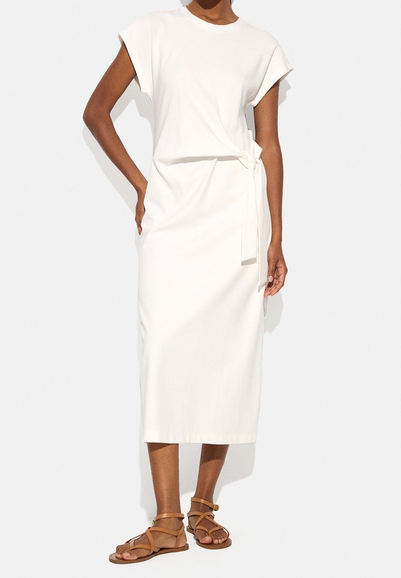 Biała midi sukienka z miękkiego materiału, z krótkimi rękawami i wiązaniem w talii. Noszona z brązowymi sandałami na paski.