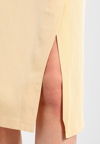 Světle žluté šaty s bočním rozparkem, vyrobené z hladké látky. Mají čisté švy a rovný lem, který zvýrazňuje nohu.