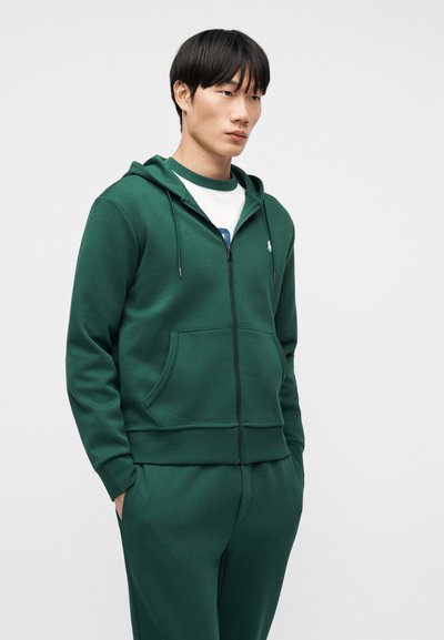 Νεαρός άνδρας με σκούρο πράσινο φούτερ με φερμουάρ και αντίστοιχο παντελόνι, στέκεται με τα χέρια στις τσέπες, κοιτάζοντας ελαφρώς στο πλάι σε απλό φόντο.