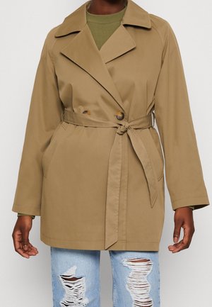 Personne portant un trench-coat beige ceinturé à la taille, un haut vert en dessous et un jean bleu clair déchiré, debout devant un fond uni.