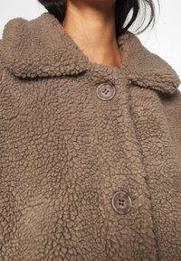 Gros plan sur une personne portant une veste polaire marron texturée avec de gros boutons marron et un large col.