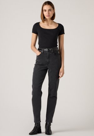 Jeune femme debout portant un haut noir à manches courtes, un jean noir taille haute avec ceinture, et des bottines noires devant un fond uni.