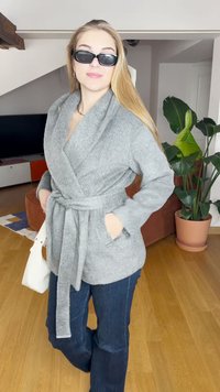 Sweater cinzento texturizado com bolsos e cinto de laço, combinado com jeans azul-escuro. O cenário inclui plantas de interior e chão de madeira.