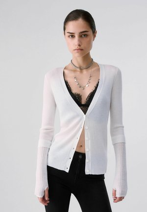 Femme portant un cardigan blanc boutonné par-dessus un bralette en dentelle noire, un pantalon noir et des colliers en argent superposés, debout devant un fond uni.
