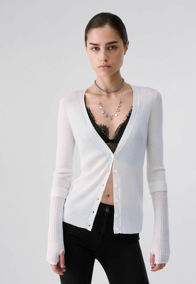Femme portant un cardigan blanc boutonné par-dessus un bralette en dentelle noire, un pantalon noir et des colliers en argent superposés, debout devant un fond uni.