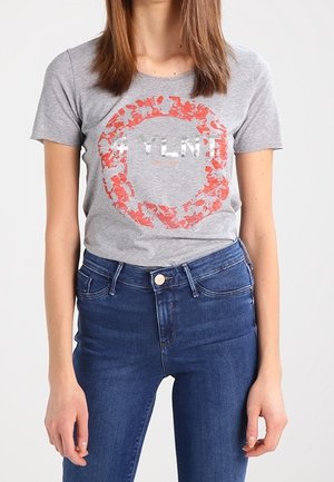 Femme portant un t-shirt gris avec un motif circulaire rouge délavé et un texte blanc, associé à un jean bleu taille haute.