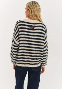 Sweter w paski w kolorze kremowym i granatowym, wykonany z miękkiego, puszystego materiału. Posiada luźny krój i metkę na górnej części pleców.