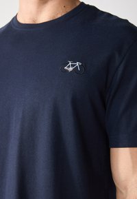 T-shirt en coton marine avec un petit motif de vélo brodé en blanc et beige dans la zone de la poitrine gauche. Manches courtes, col rond.