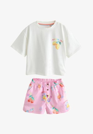 Gyerek fehér, túlméretezett pólót visel, nyári témájú mintákkal, valamint rózsaszín rövidnadrágot jégkrém és desszert mintákkal, háttal áll.