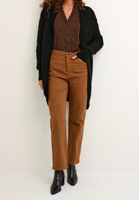 Kvinna i svart kofta, mönstrad brun blus, bruna byxor och svarta klackade ankelboots mot en enfärgad bakgrund.