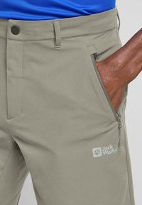 Pantalons d'extérieur gris clair avec une poche zippée, fermeture à bouton et marquage. Fabriqués à partir d'un tissu lisse et texturé.