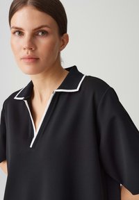 Schwarzes Poloshirt mit weißem Kragen in kleiner Größe und dezentem Halsreißverschluss. Der Stoff wirkt glatt und hat eine lockere Passform sowie kurze Ärmel.