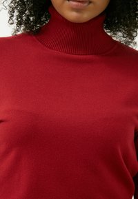 Roter Rollkragenpullover aus Strickstoff. Mit einem gerippten Kragen und einer glatten, strukturierten Oberfläche. Nahaufnahme, die die Stoffdetails hervorhebt.