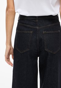 Mørk denimjeans med høy midje, to baklommer og synlig oransje søm. Stoffet har en teksturert overflate.