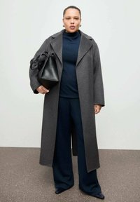 Cappotto di lana grigio con ampi revers, abbinato a un dolcevita blu scuro e pantaloni a gamba larga coordinati. Borsa in pelle nera e scarpe a punta.