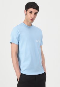 T-shirt en coton bleu clair avec un col rond et des manches courtes, arborant un petit logo blanc sur la poitrine gauche. Texture lisse.