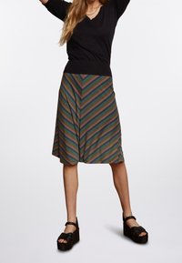 Černý pletený top s výstřihem do V, doplněný sukní s multibarevným vzorem ve tvaru šipky. Modelka má na sobě černé sandály na platformě.