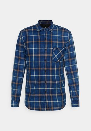 Chemise bleue à manches longues avec motif à carreaux marron et blanc et une poche poitrine unique inclinée en diagonale sur le côté droit.