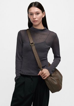 Frau mit langen dunklen Haaren trägt ein durchscheinendes, dunkelgraues Langarmshirt, lockere schwarze Hose und eine braune Lederschultertasche mit breitem Riemen.