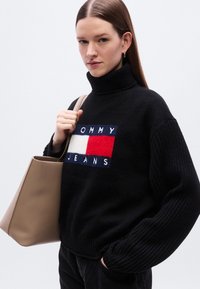 Pull à col roulé noir en maille avec un patch logo brodé coloré. Le modèle tient un sac fourre-tout beige, avec un arrière-plan minimaliste.