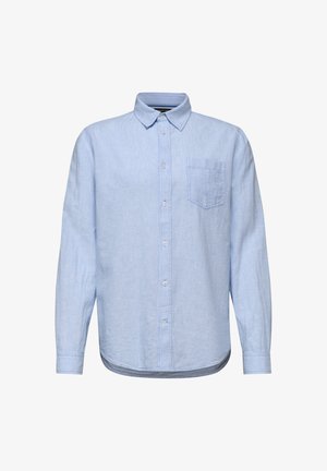 Man in lichtblauw overhemd met knopen en borstzak, die naar links kijkt tegen een neutrale achtergrond.