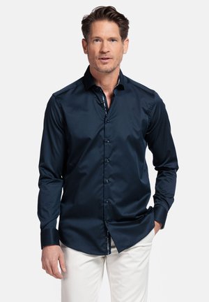 Chemise à manches longues bleu marine en tissu lisse avec un boutonnage à l'avant et un détail de col intérieur contrastant, associée à un pantalon clair.