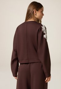 Bruine sweatshirt met lange mouwen en geribbelde onderrand, voorzien van ingewikkelde witte bloemenborduursels op de linkerschouder. Soepele textuur.