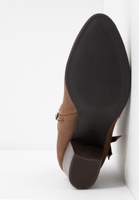 Brązowy zamszowy but z gumową podeszwą, zaokrąglonym noskiem, dekoracyjnym szyciem i bocznym zamkiem błyskawicznym. Płaski obcas z teksturowanym dnem.