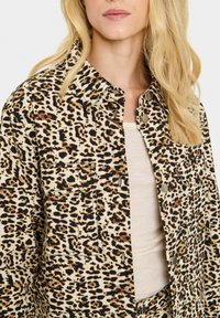 Jakna s leopard uzorkom napravljena od tkanine s bež podlogom i crnim/smeškim mrljama; ima klasičan ovratnik i prednje džepove sa dugmadi.