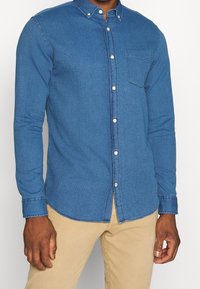 Blå chambray-skjorta med knappar, långa ärmar, krage, bröstficka och kontrasterande knappar; strukturerad tyg med en lätt mönstring.
