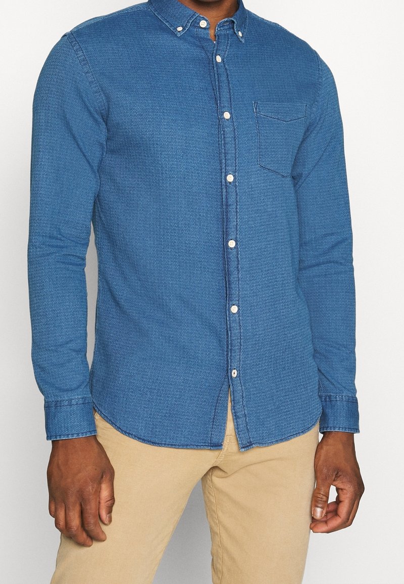 Blå chambray-skjorta med knappar, långa ärmar, krage, bröstficka och kontrasterande knappar; strukturerad tyg med en lätt mönstring.