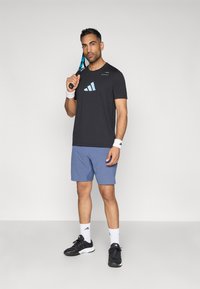 Melna sporta T-krekls ar gaiši zilā krāsā logo, apvienots ar ziliem šortiem. Aksesuāri ietver baltus aproces un melnas sporta kurpes.