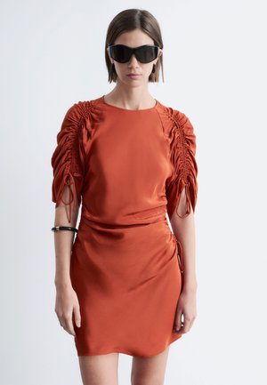 Kobieta w czarnych, dużych okularach przeciwsłonecznych oraz rdzawej, pomarańczowej sukience z marszczonego satyny z rękawami do połowy ramienia i czarną bransoletką na lewej ręce.